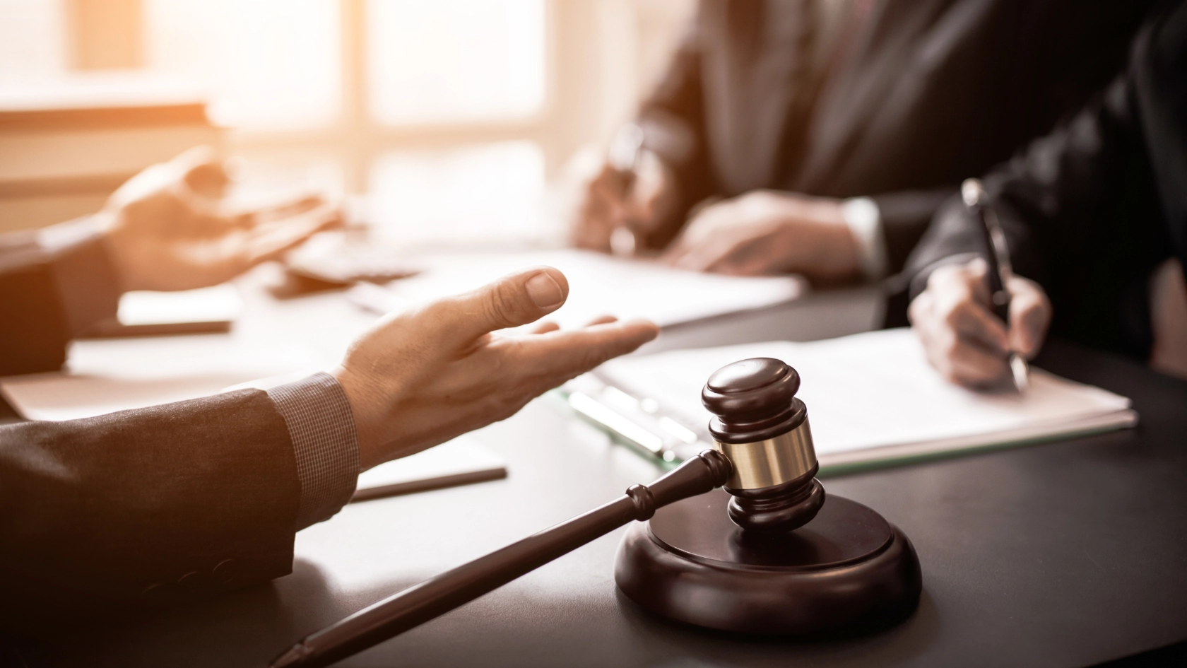 Drei Männer diskutieren über Recht an einem Schreibtisch, Akten und Richterhammer sind zu sehen, ein Mann notiert sich etwas