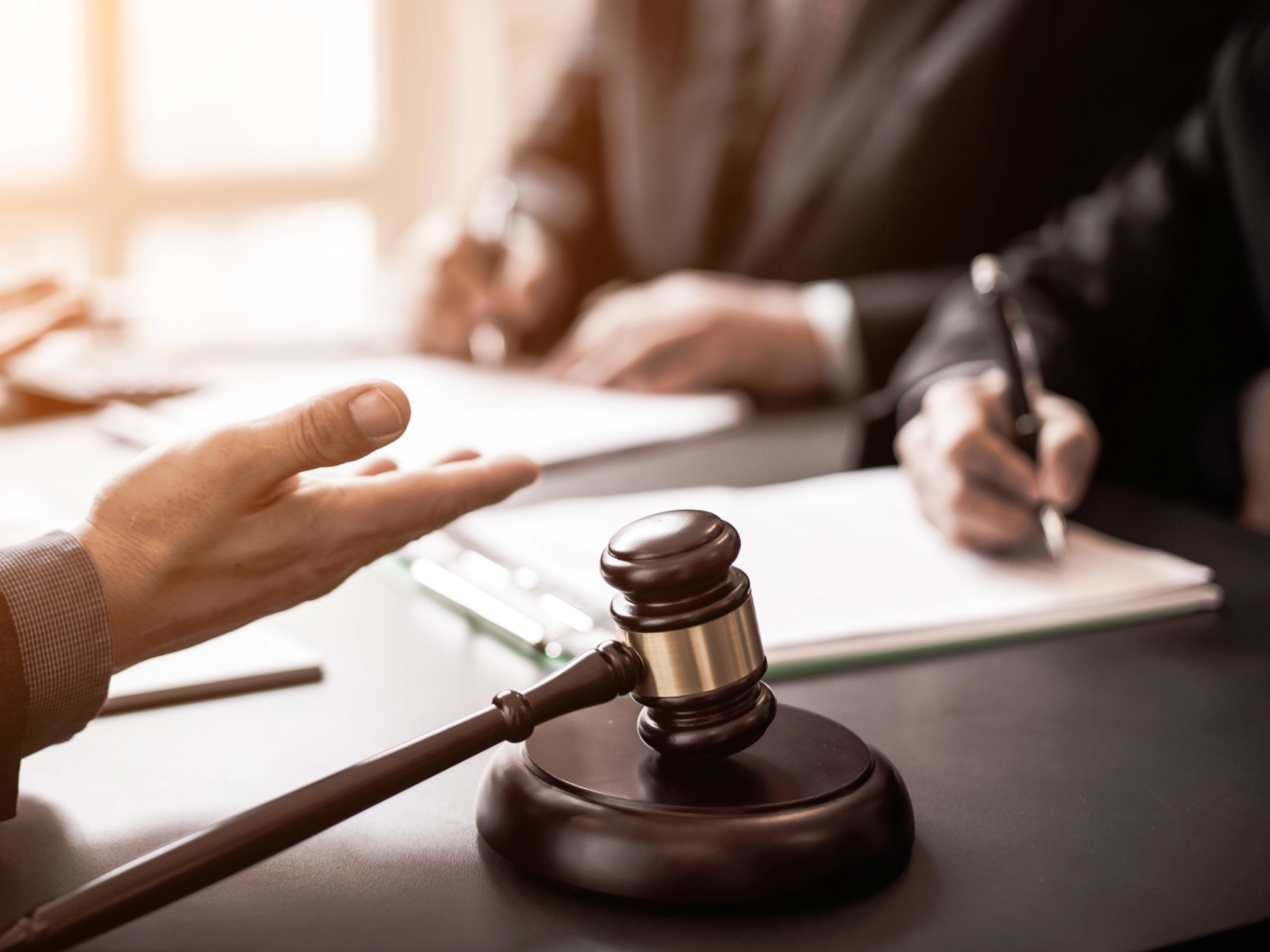 Drei Männer diskutieren über Recht an einem Schreibtisch, Akten und Richterhammer sind zu sehen, ein Mann notiert sich etwas